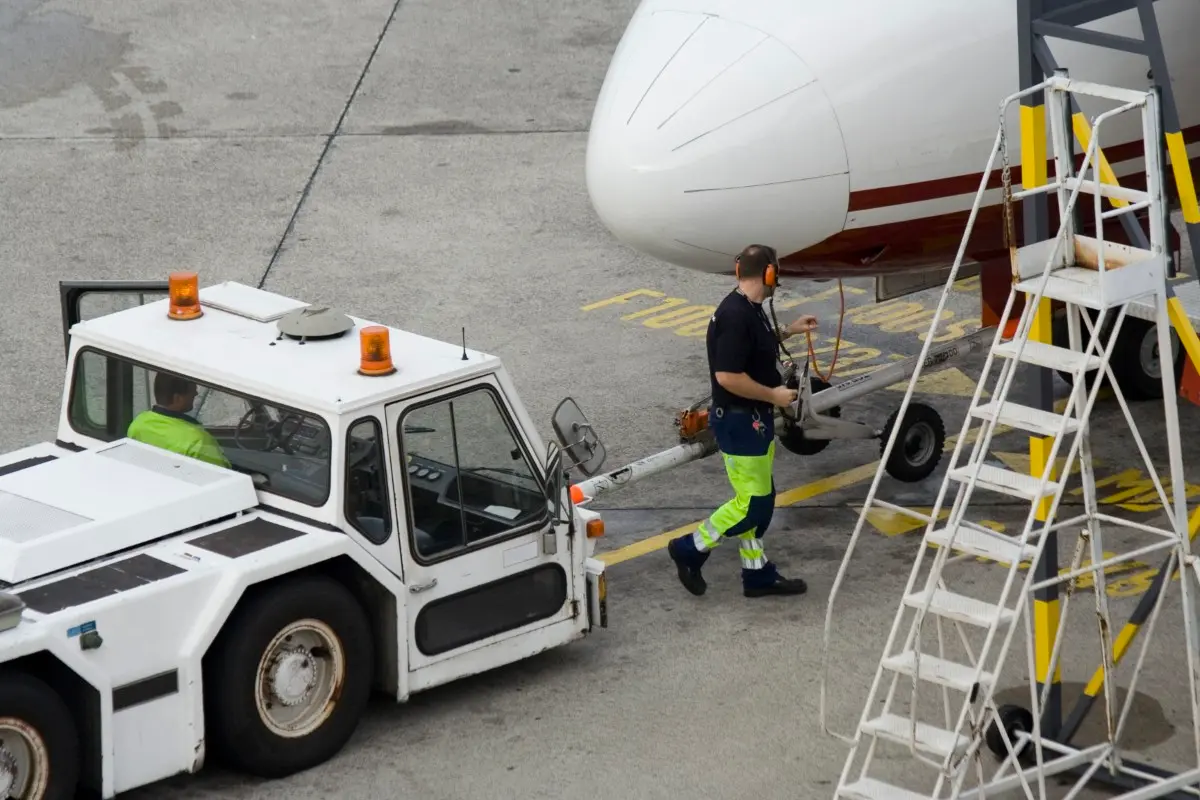 Airport ground crew preparing a plane for departure. A worker in high-visibility clothing walks toward the aircraft, while another operates a tug vehicle nearby.
