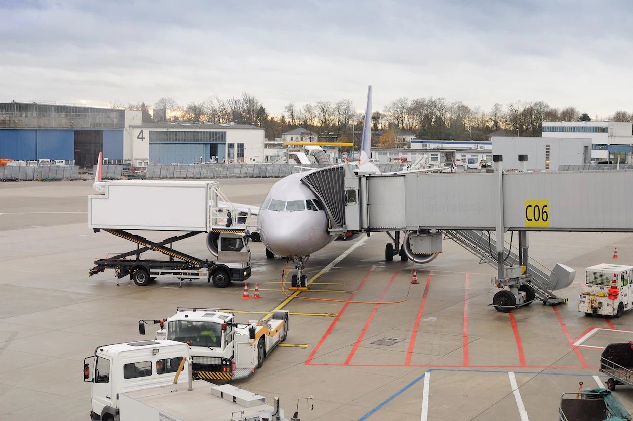 Airplane at gate C06 with jet bridge attached. Ground crew vehicles and equipment surround the plane on a cloudy day at the airport.