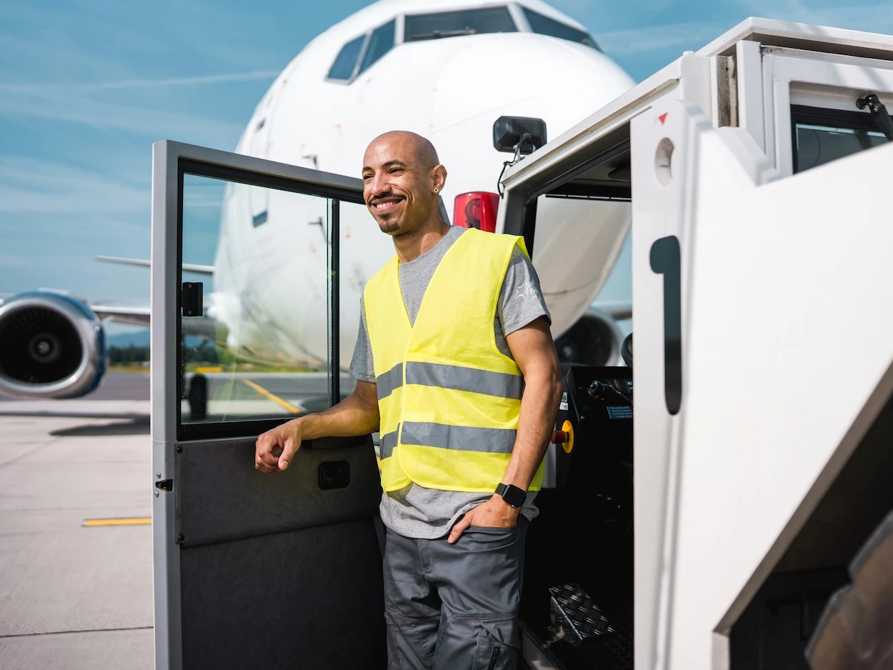 Smiling airport worker in a yellow vest stands by an open vehicle door on the tarmac, with a large aircraft in the background under a clear sky.