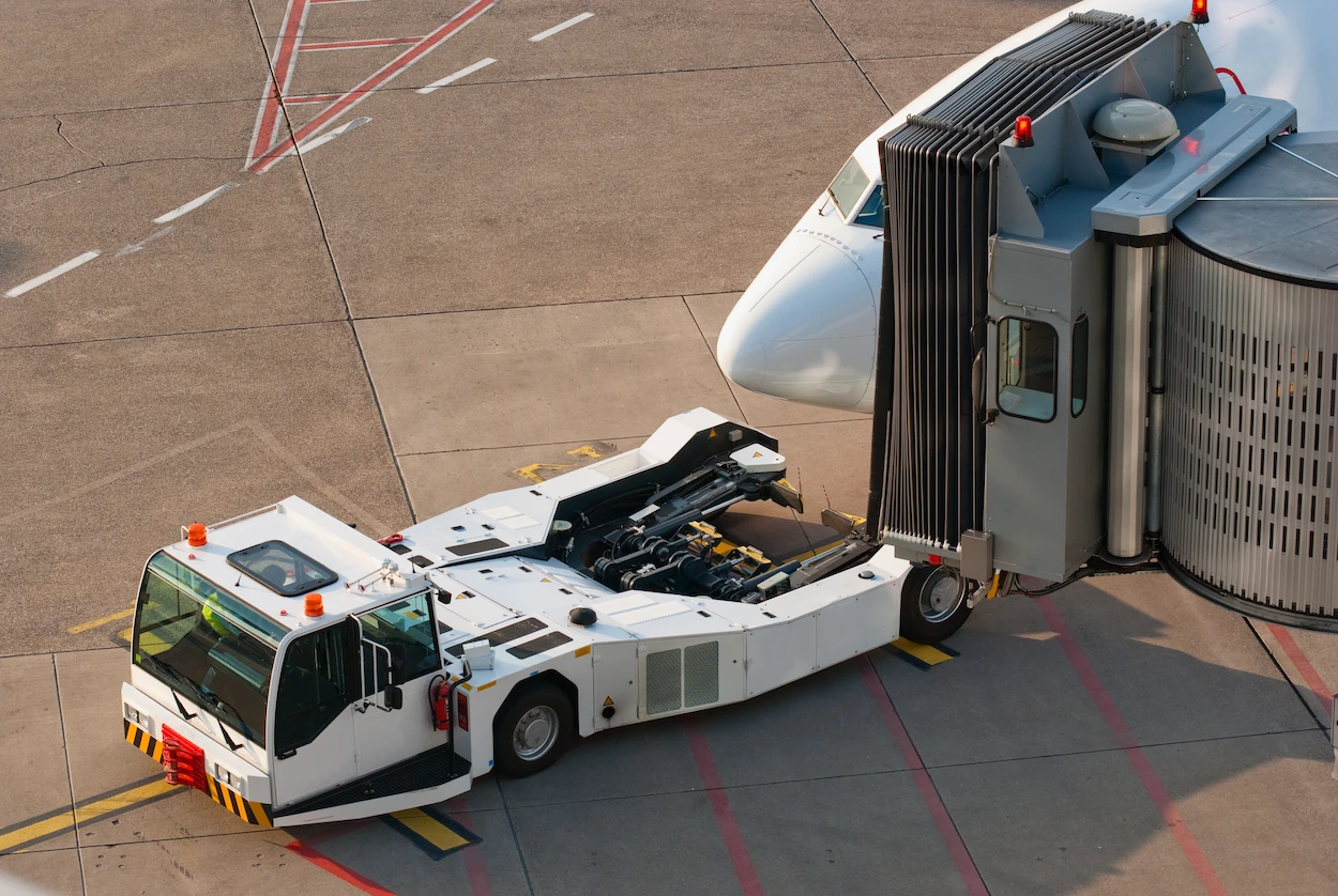 Airport scene showing an airplane nose docked at a jet bridge with a white aircraft tug attached. The area is marked by yellow and red lines.