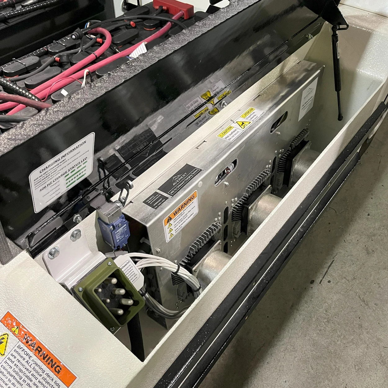 Open electrical panel with exposed components, including wires and circuit boxes, featuring warning labels and visible connectors, in a workshop setting.