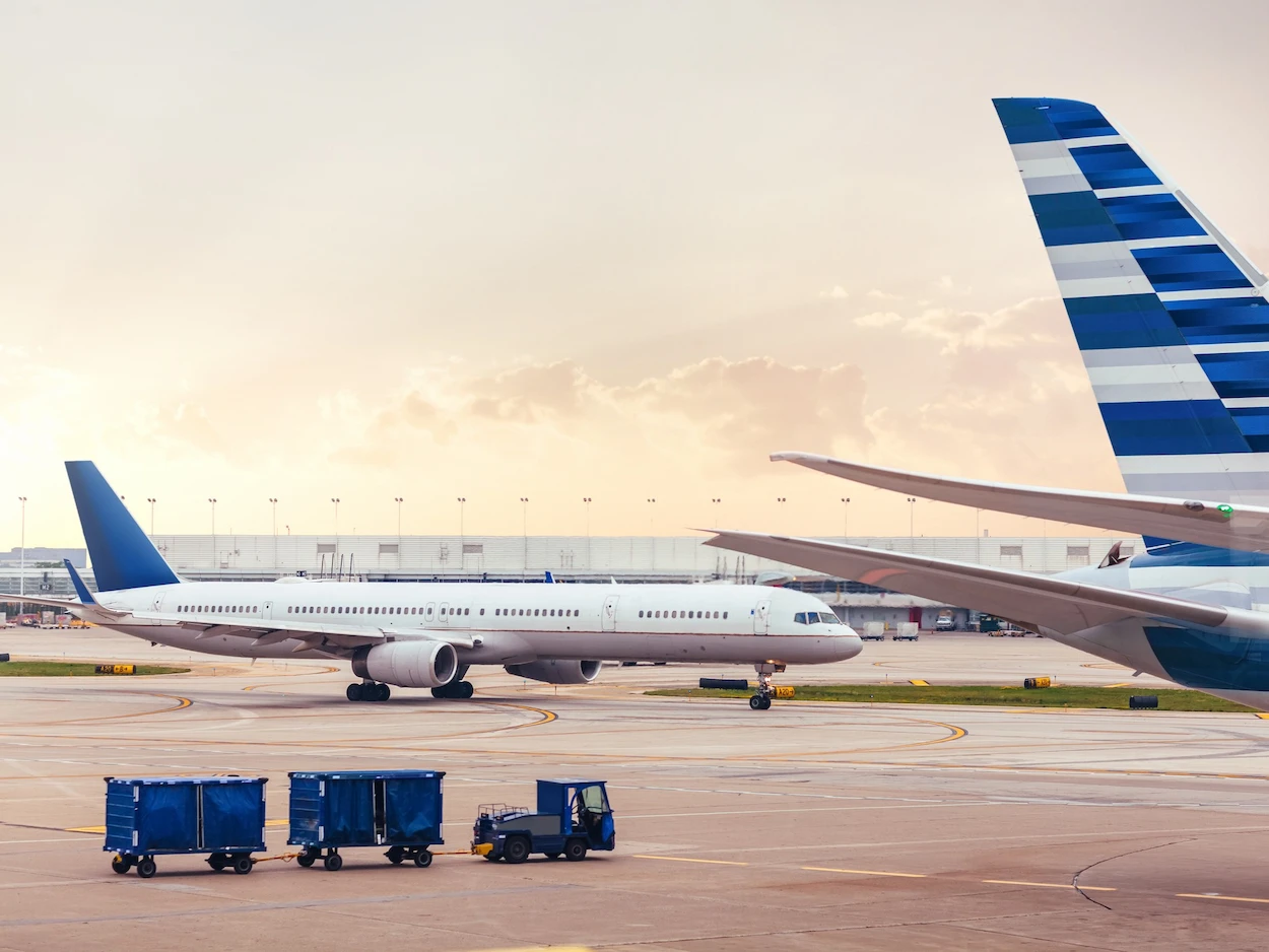 A large passenger airplane on a runway during sunset, with another plane's tail visible in the foreground. A luggage cart sits to the left, adding a sense of airport activity.