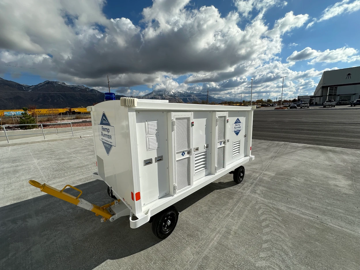 White utility trailer with "Ramp Runner" logo stands on tarmac under a cloudy sky, with snow-capped mountains and an industrial building in the background.