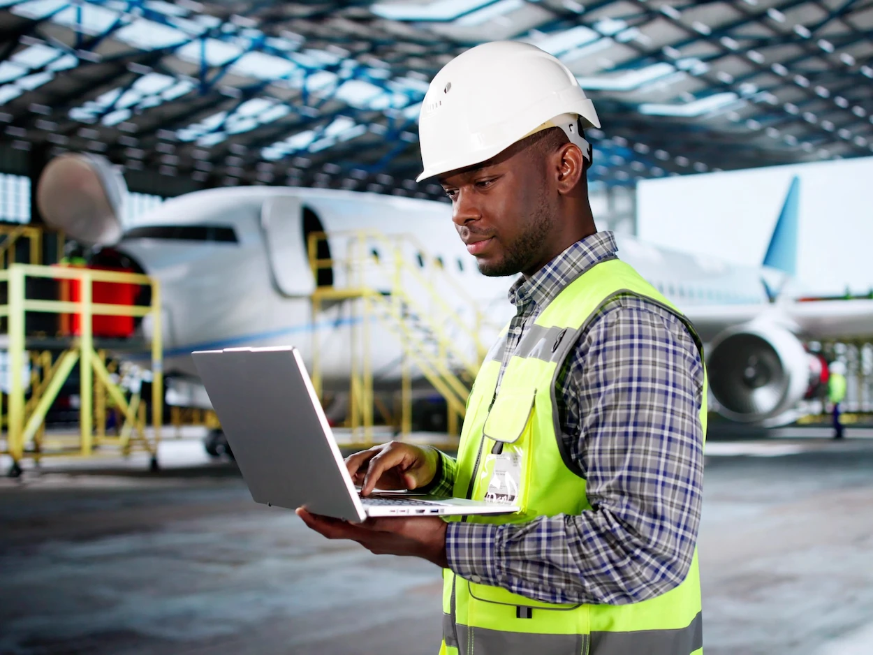 A man in a reflective vest and hard hat uses a laptop, standing in an aircraft hangar with a plane in the background. The setting is professional and focused.