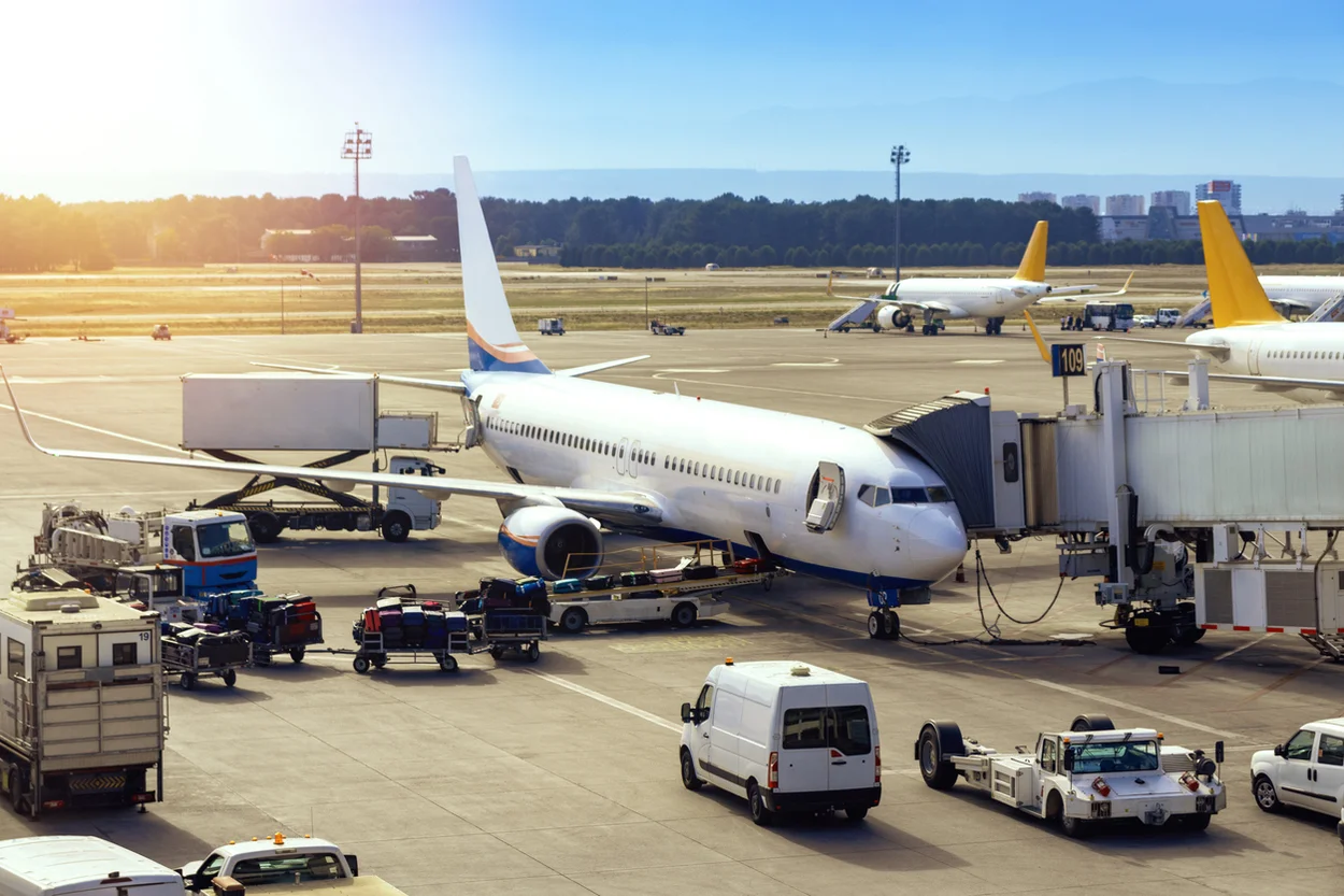 A passenger airplane parked at a jet bridge on a busy airport ramp, surrounded by ground support vehicles, baggage carts, and service vans.