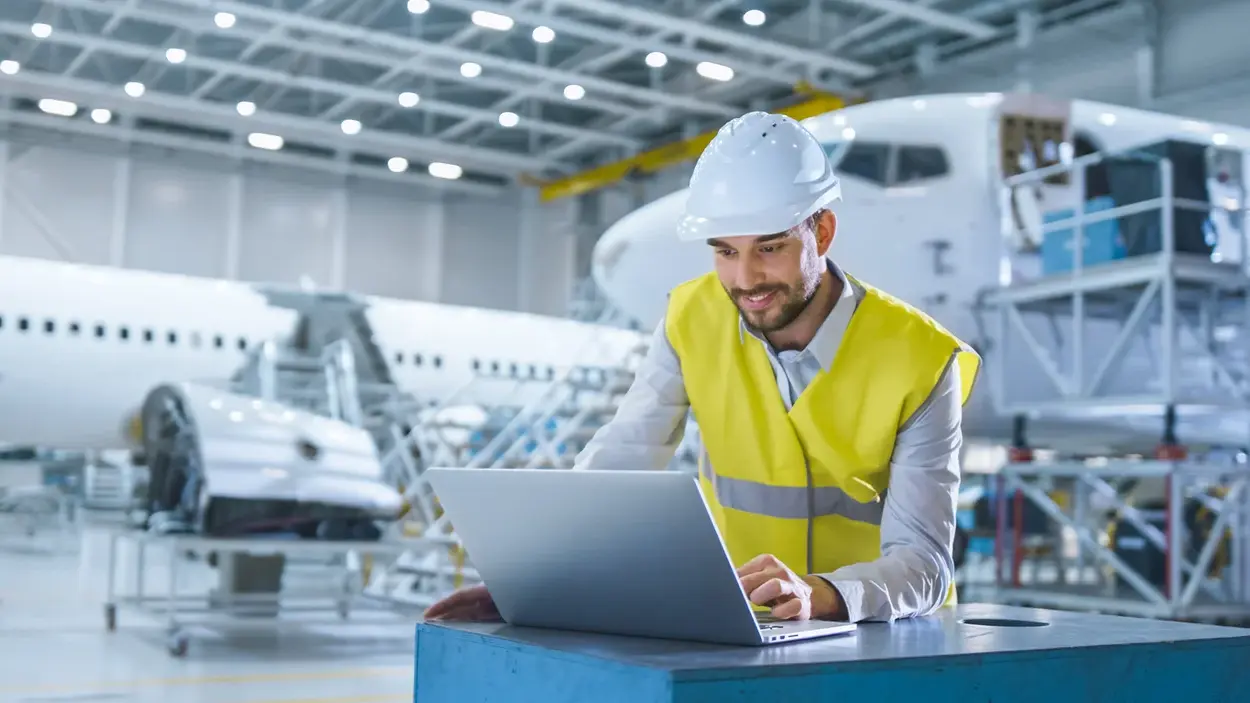 An engineer in a hard hat and safety vest works on a laptop inside an aircraft maintenance hangar, with an airplane in the background.